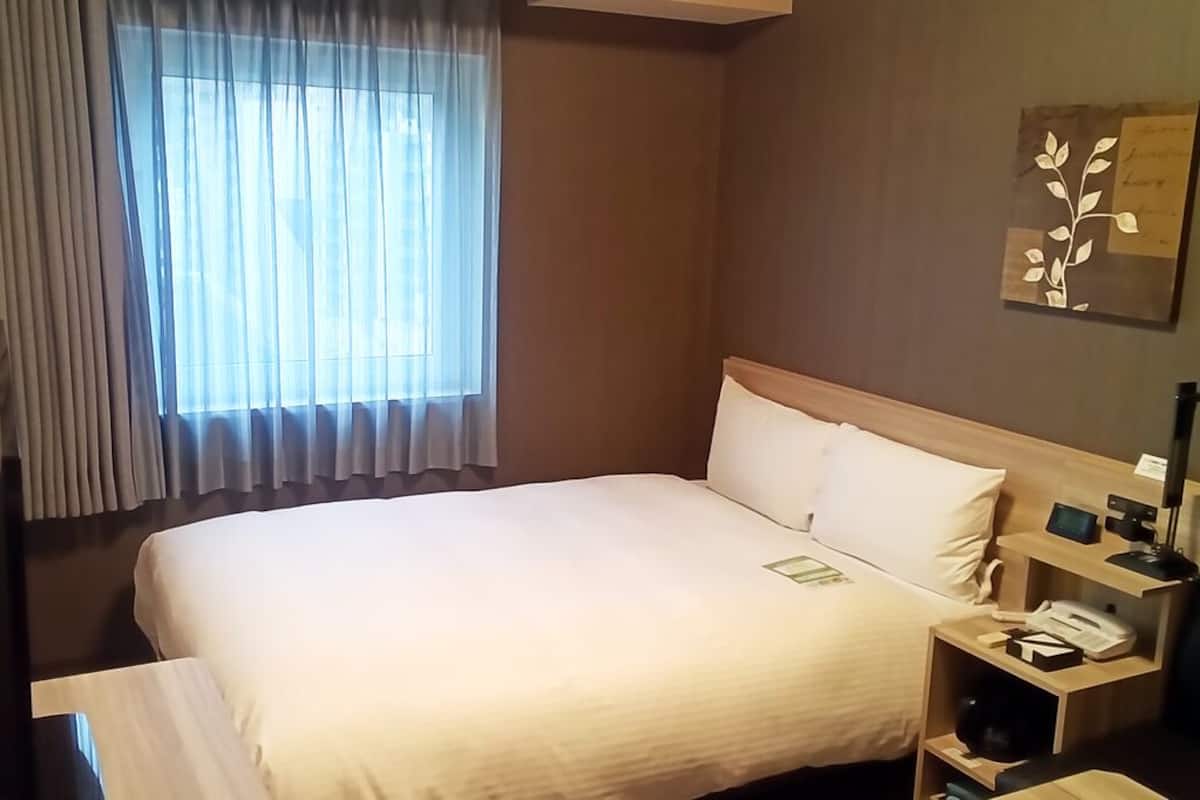 A hotel room with a bed, a nightstand with a lamp, a telephone, and a window with curtains.