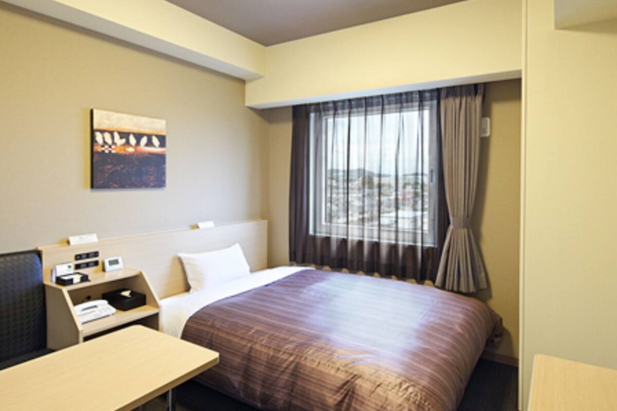 A hotel room with a bed, a desk, a chair, a window with curtains, and a painting on the wall.