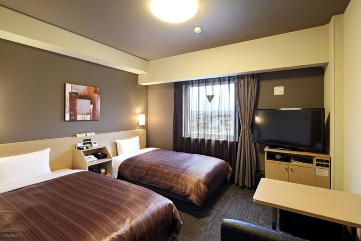 A hotel room with two beds, a TV, a desk, and a window with curtains.