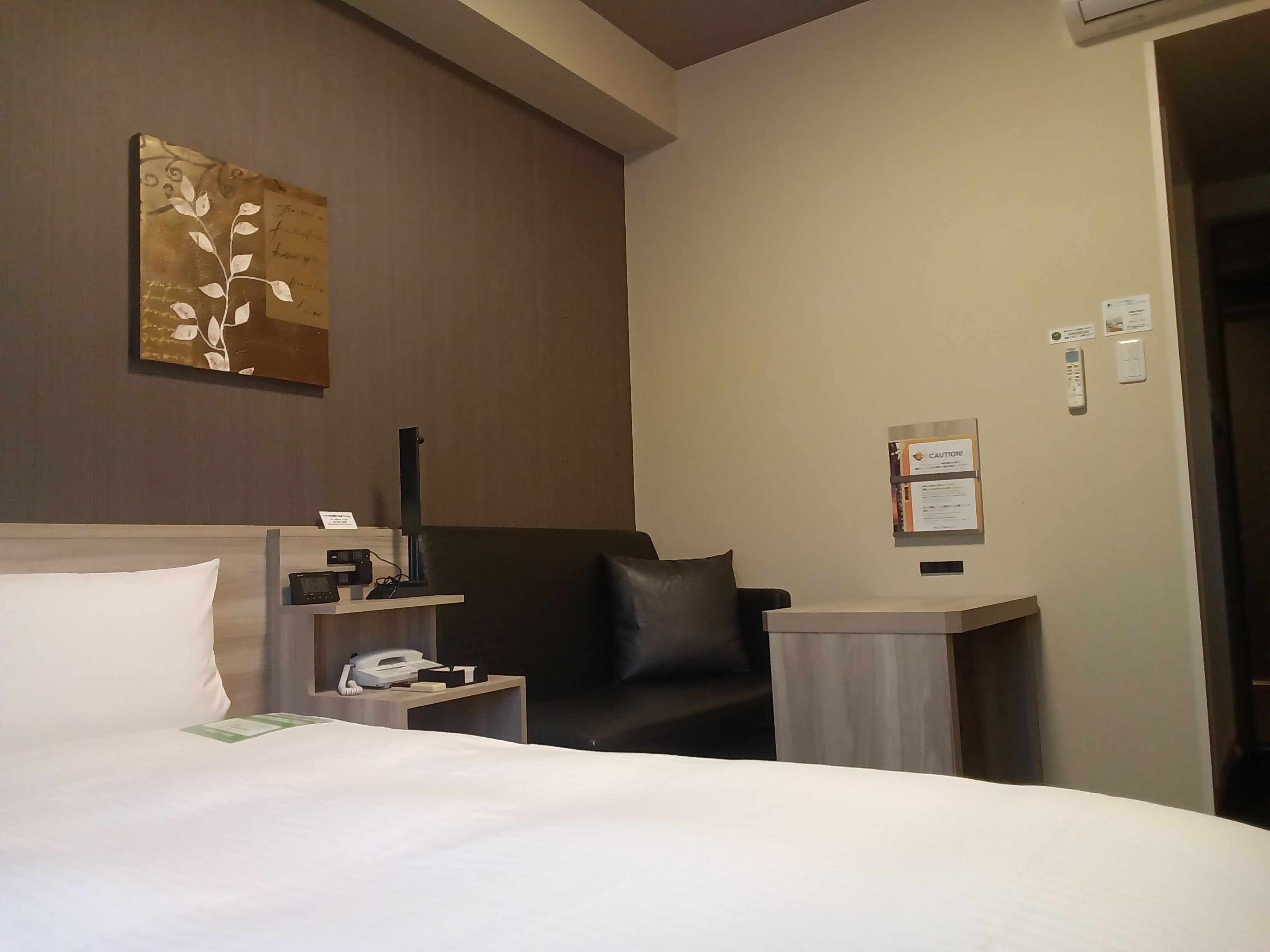 A hotel room with a bed, a desk, a chair, a telephone, and a wall-mounted control panel.