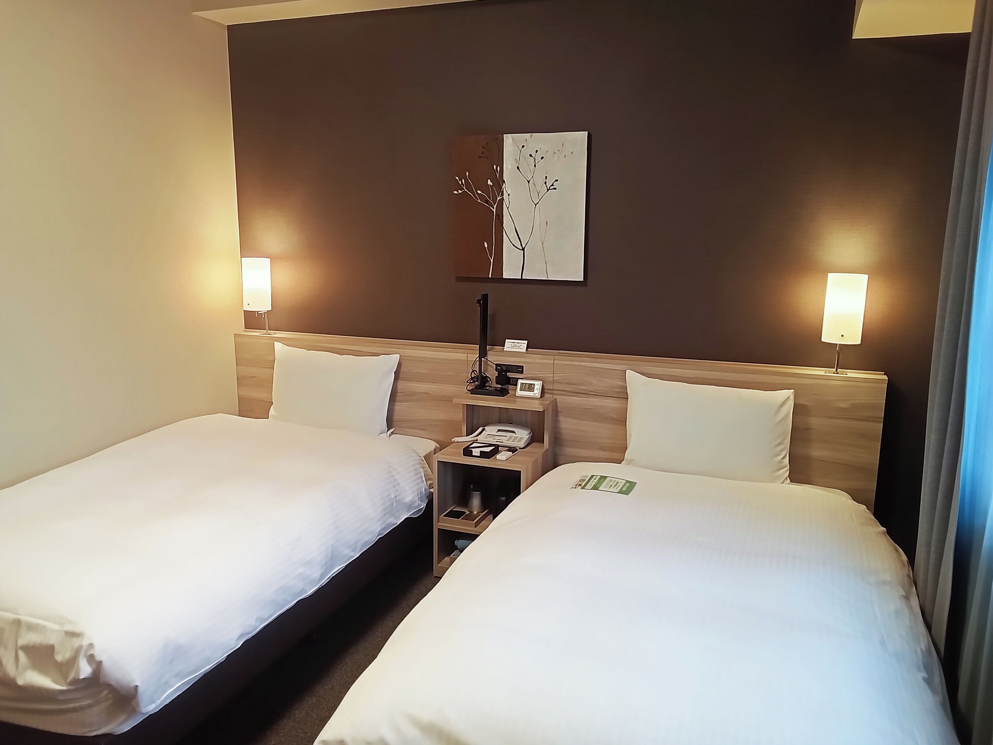 A hotel room with two single beds, a nightstand with a phone and a lamp, and a wall-mounted artwork.