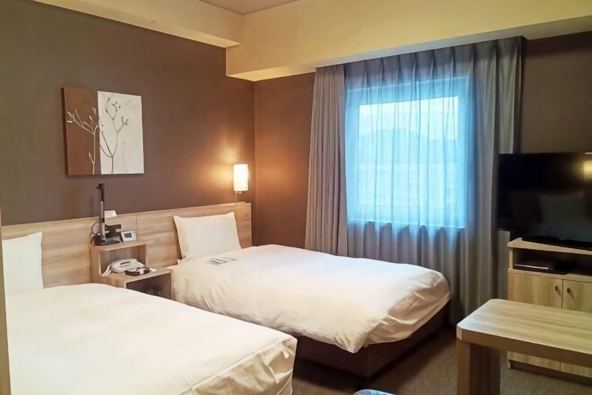 A hotel room with two beds, a TV, a desk, and a window with curtains.