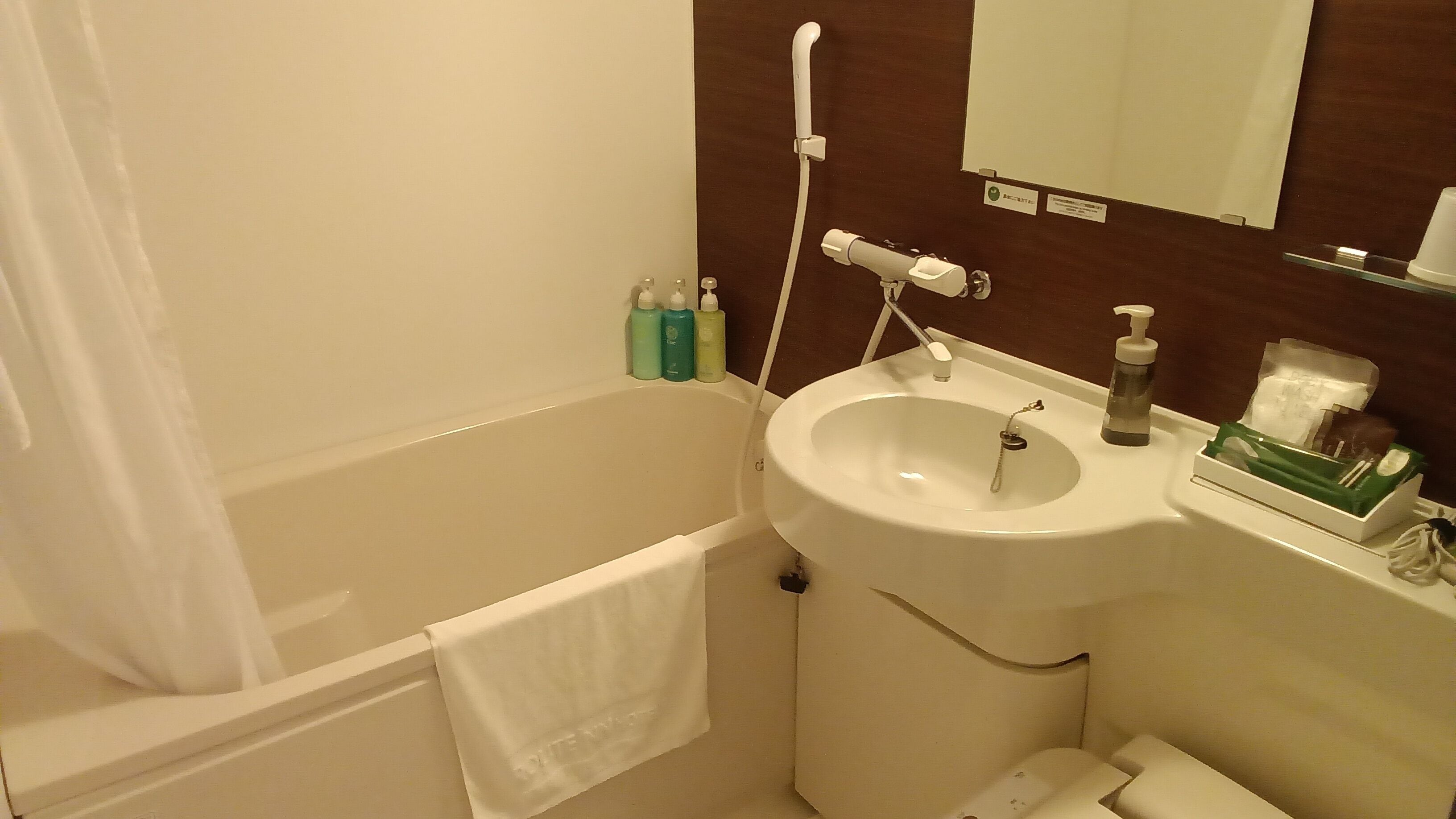 A bathroom with a bathtub, sink, and mirror. There are toiletries on the sink and a towel hanging on the wall.