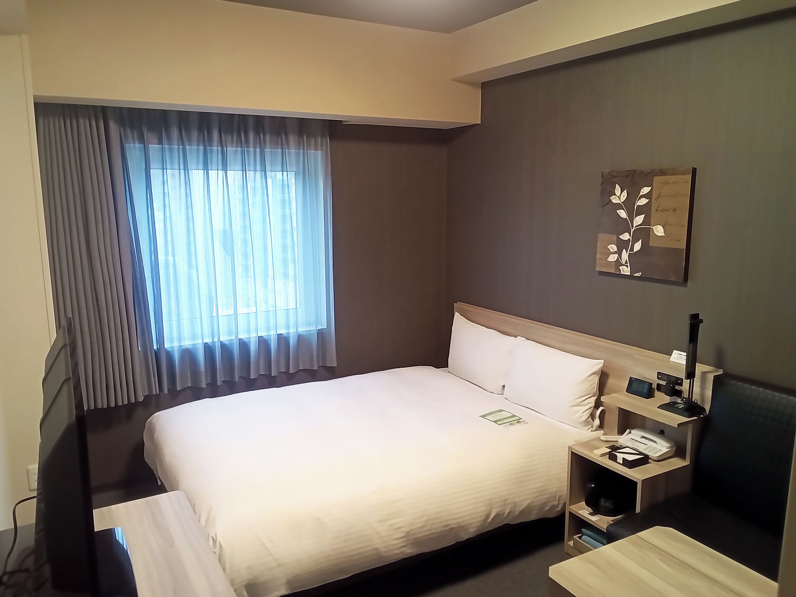 A hotel room with a bed, bedside table, lamp, and a window with curtains.