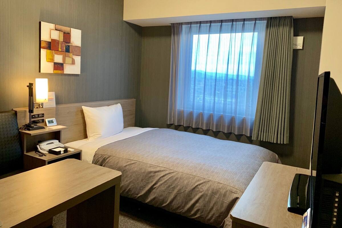 A hotel room with a bed, a desk, a telephone, a television, and a window with curtains.