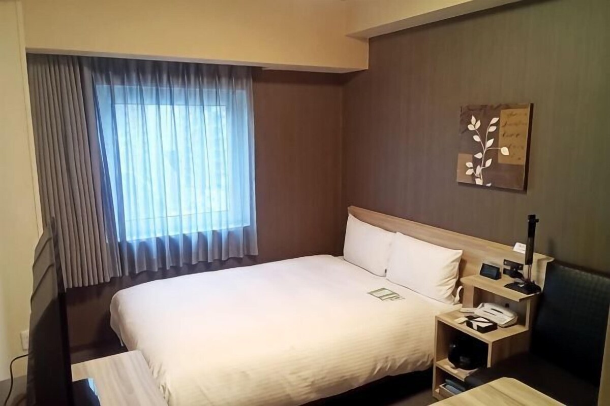 A hotel room with a bed, a bedside table with a lamp, a nightstand, and a window with curtains.