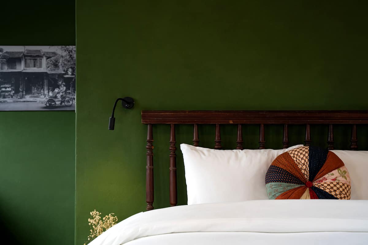 A bed with white bedding, a wooden headboard, and a colorful pillow. A black lamp is on the wall to the left.