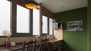 A kitchen with a wooden table, chairs, a hanging light fixture, a window with curtains, a green wall, and a mounted television. - Centraltique Hidden Oasis (Hanoi)
