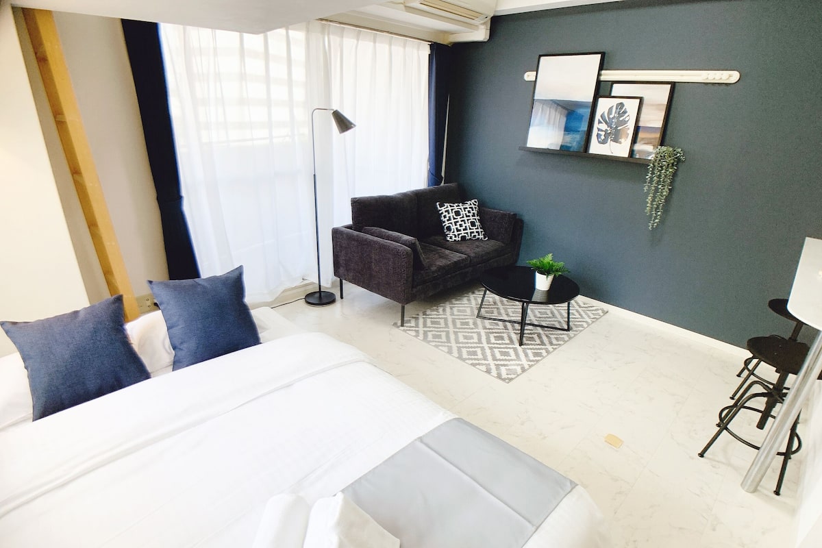 A modern hotel room with a large bed, a dark armchair, a small round table with a plant, and a desk with a chair.