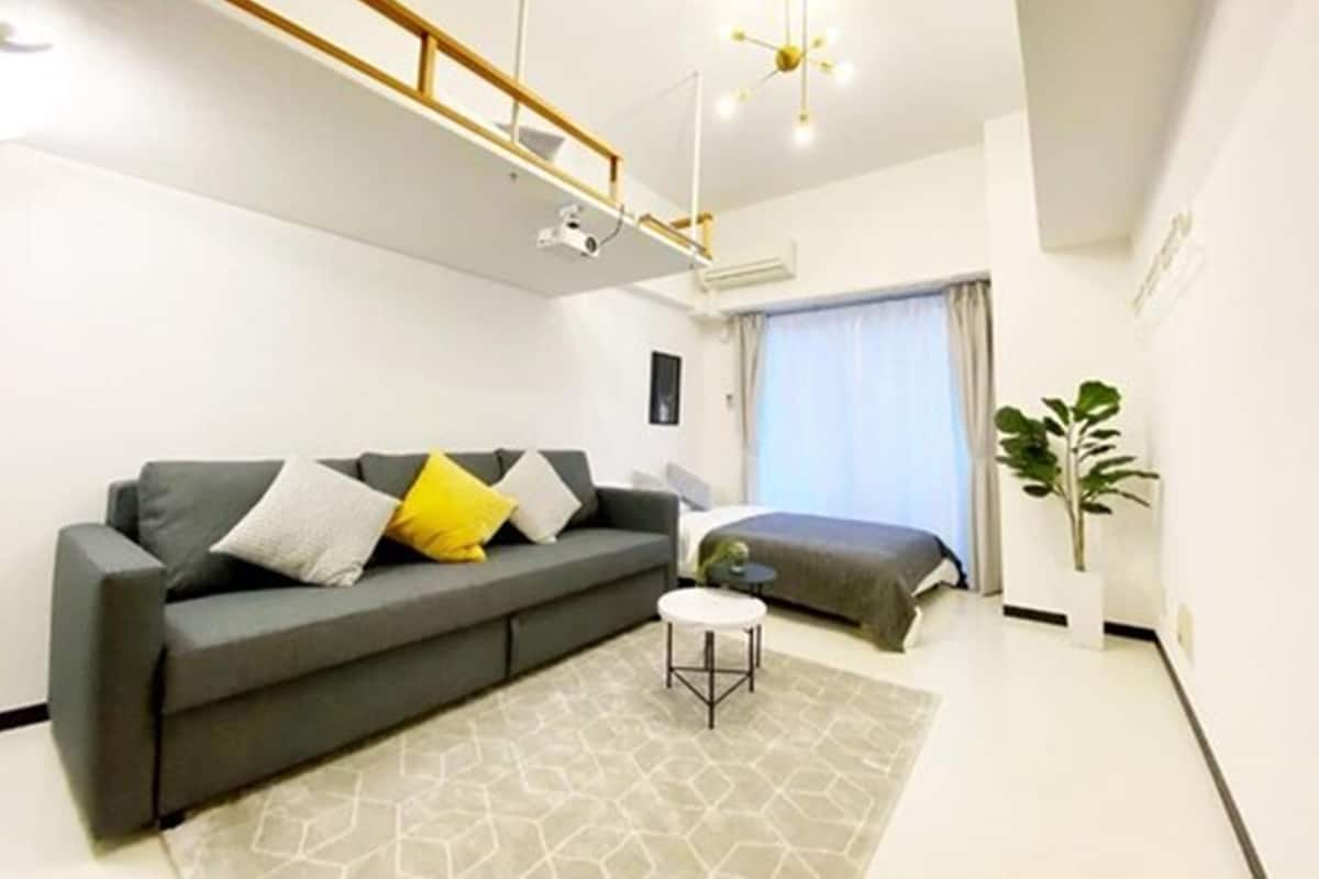 Ein modernes Wohnzimmer mit einem grauen Sofa, einem kleinen runden Tisch, einem Bett und einem großen Fenster mit Vorhängen.