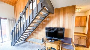 A living room with a wooden staircase, a flat-screen TV, a wooden bench, and a kitchen area in the background.