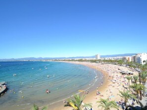 A beach with clear blue water, a sandy shore, and a row of buildings along the coastline.