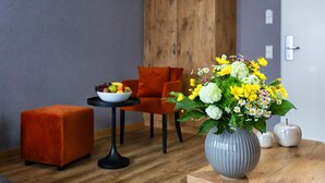Una sala de estar moderna con piso de madera, un florero gris con flores amarillas, una mesa redonda con un cuenco de frutas y un sillón naranja.