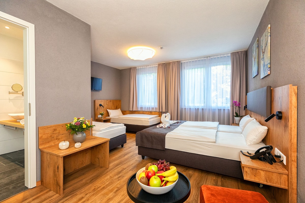 A hotel room with two beds, a wooden desk, a bowl of fruit, and a bathroom visible through an open door.