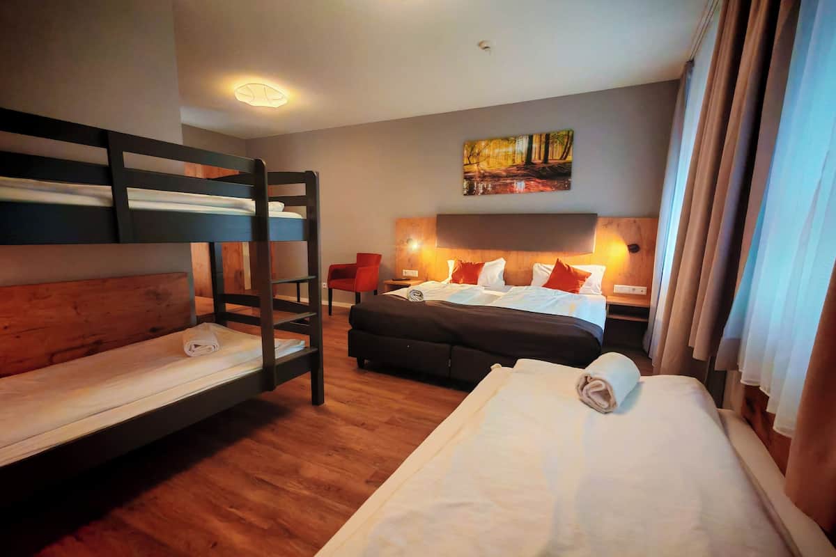 A hotel room with bunk beds, a single bed, a wooden headboard, a red chair, a painting on the wall, and a window with curtains.
