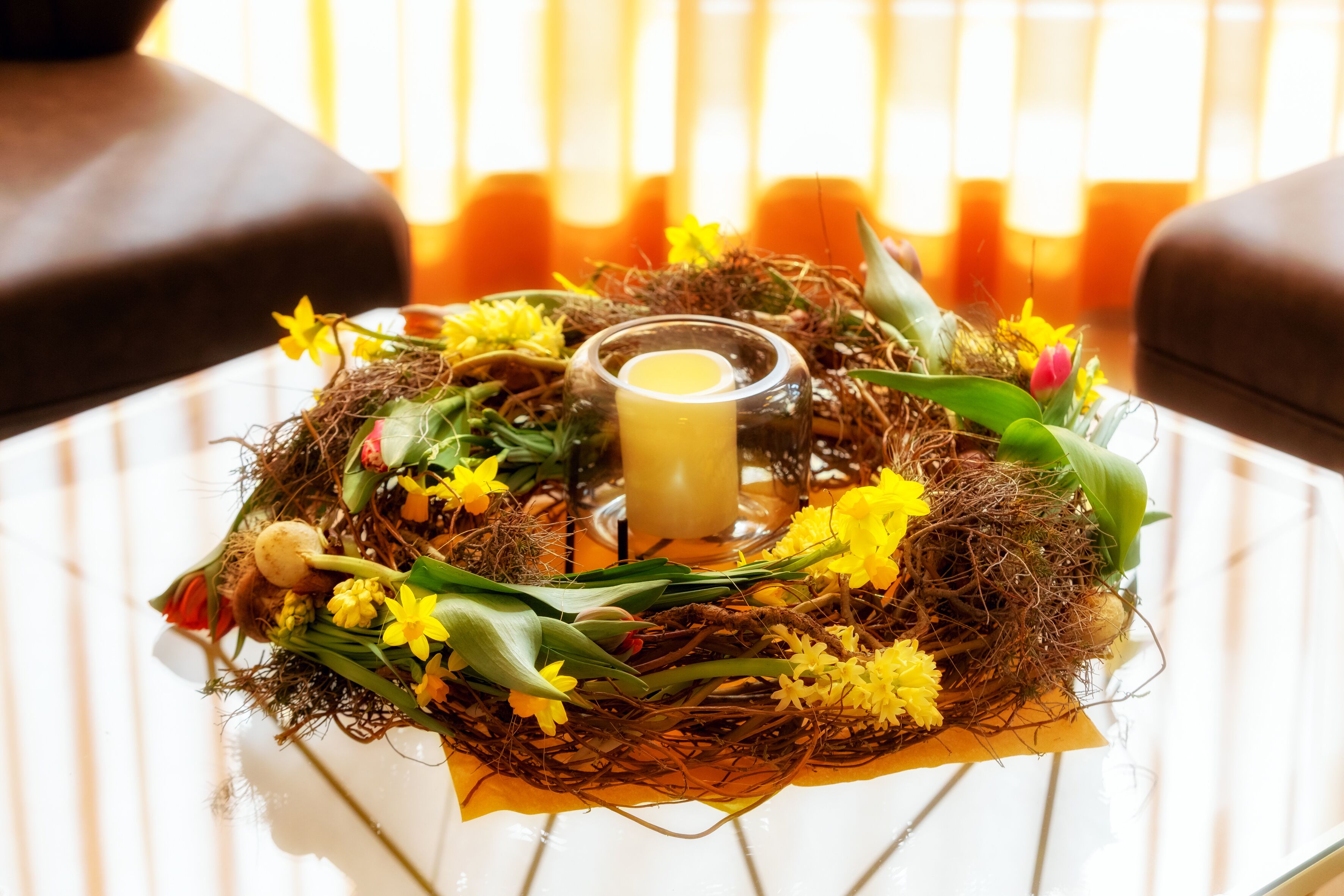 A decorative arrangement on a table, including a glass candle holder, a nest-like structure with flowers and greenery, and a candle.