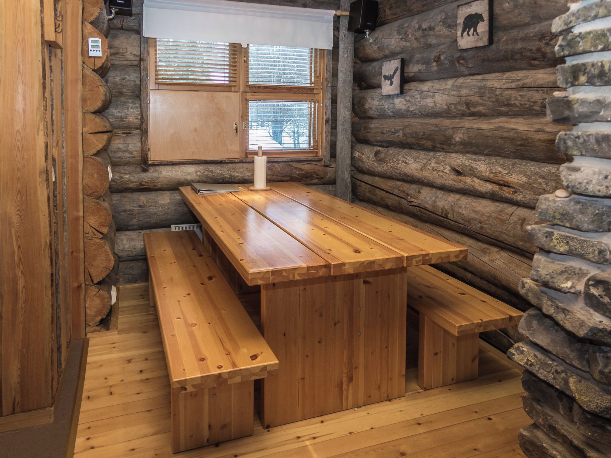 A wooden log cabin interior with a central wooden table and benches, a window with blinds, and a wall with animal silhouettes.