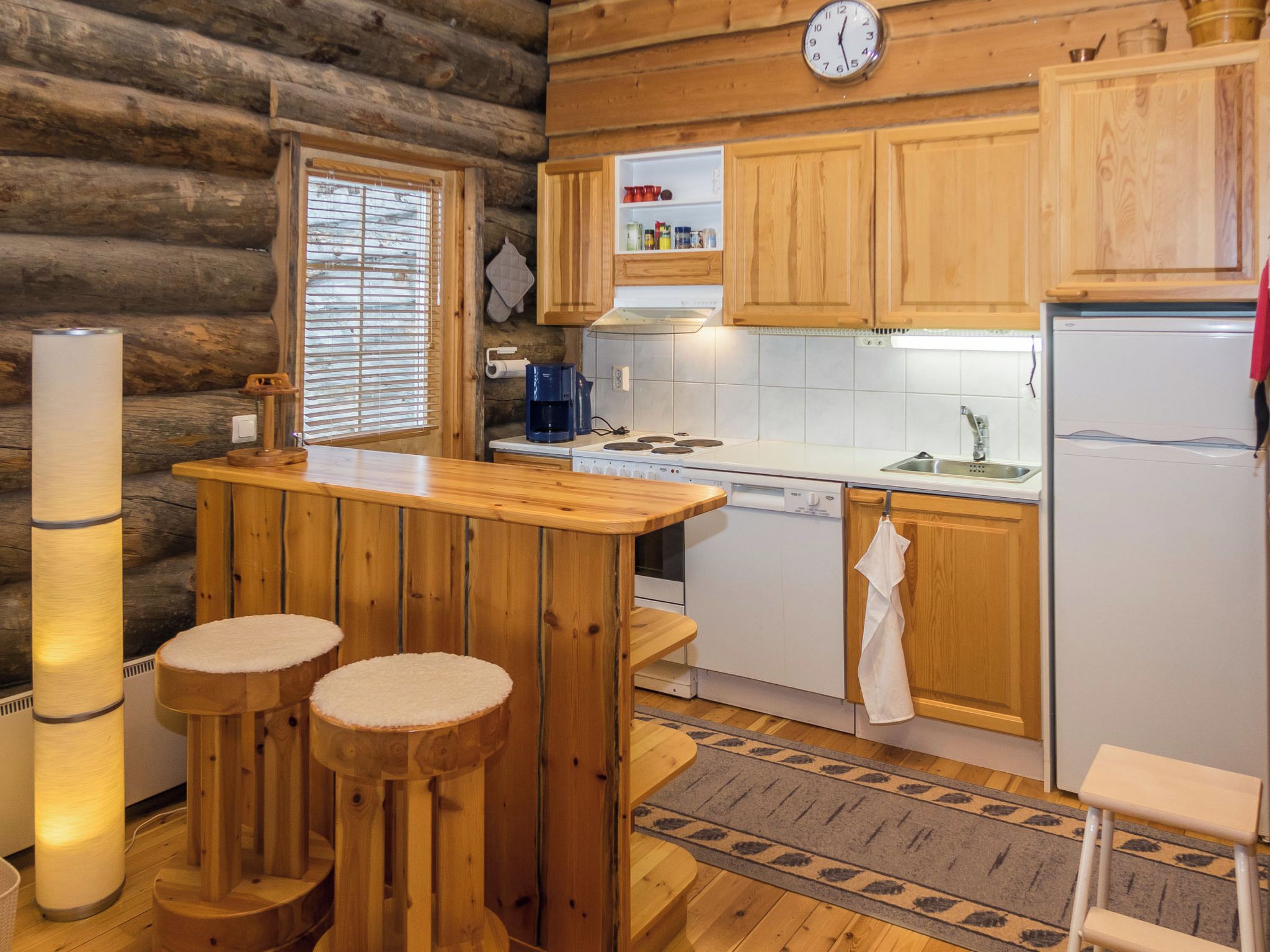 A cabin-style kitchen with wooden cabinets, a stove, a sink, and a refrigerator. There are stools at the bar and a window with blinds.