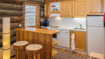 A cabin-style kitchen with wooden cabinets, a stove, a sink, and a refrigerator. There are stools at the bar and a window with blinds.