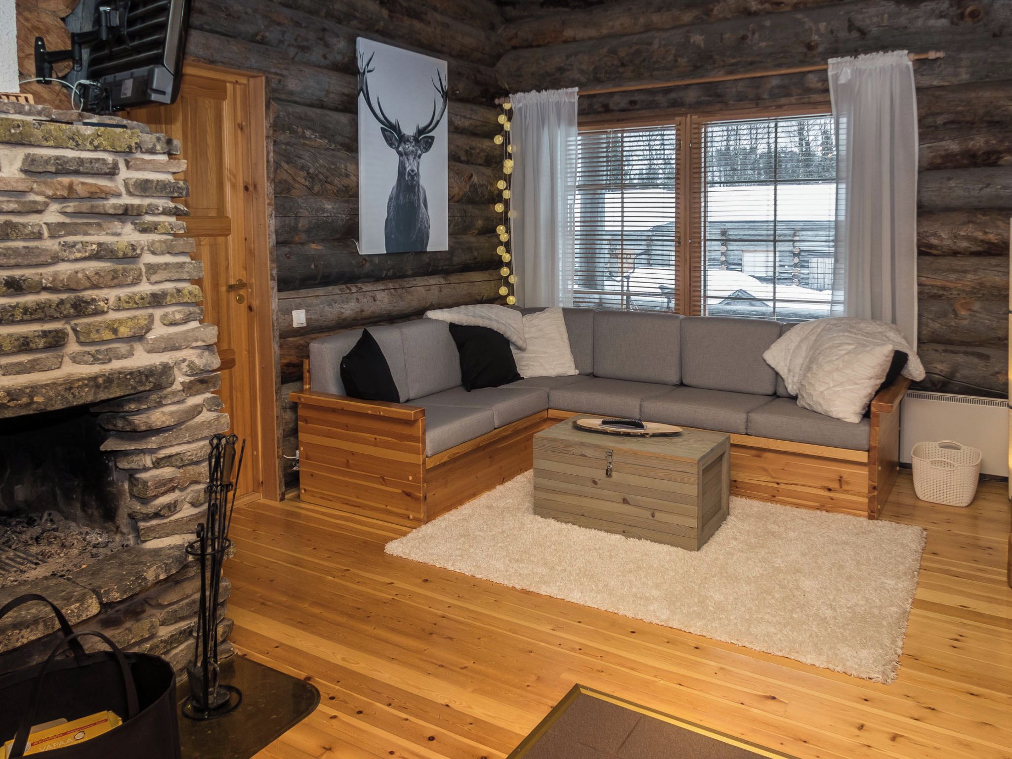 L’intérieur d’un chalet confortable, avec des murs en rondins de bois, une cheminée en pierre, un canapé en bois et une grande fenêtre équipée de stores.