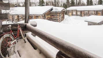 A wooden log cabin with a snow-covered roof, a bicycle leaning against a wooden post, and a snowy landscape with trees in the background.