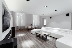 A modern living room with a white sofa, a coffee table, and a TV. There is a dining area with a table and chairs.