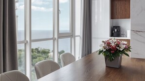 Un espace repas moderne avec une table et des chaises en bois, un arrangement floral et une vue sur la mer grâce à de grandes fenêtres.