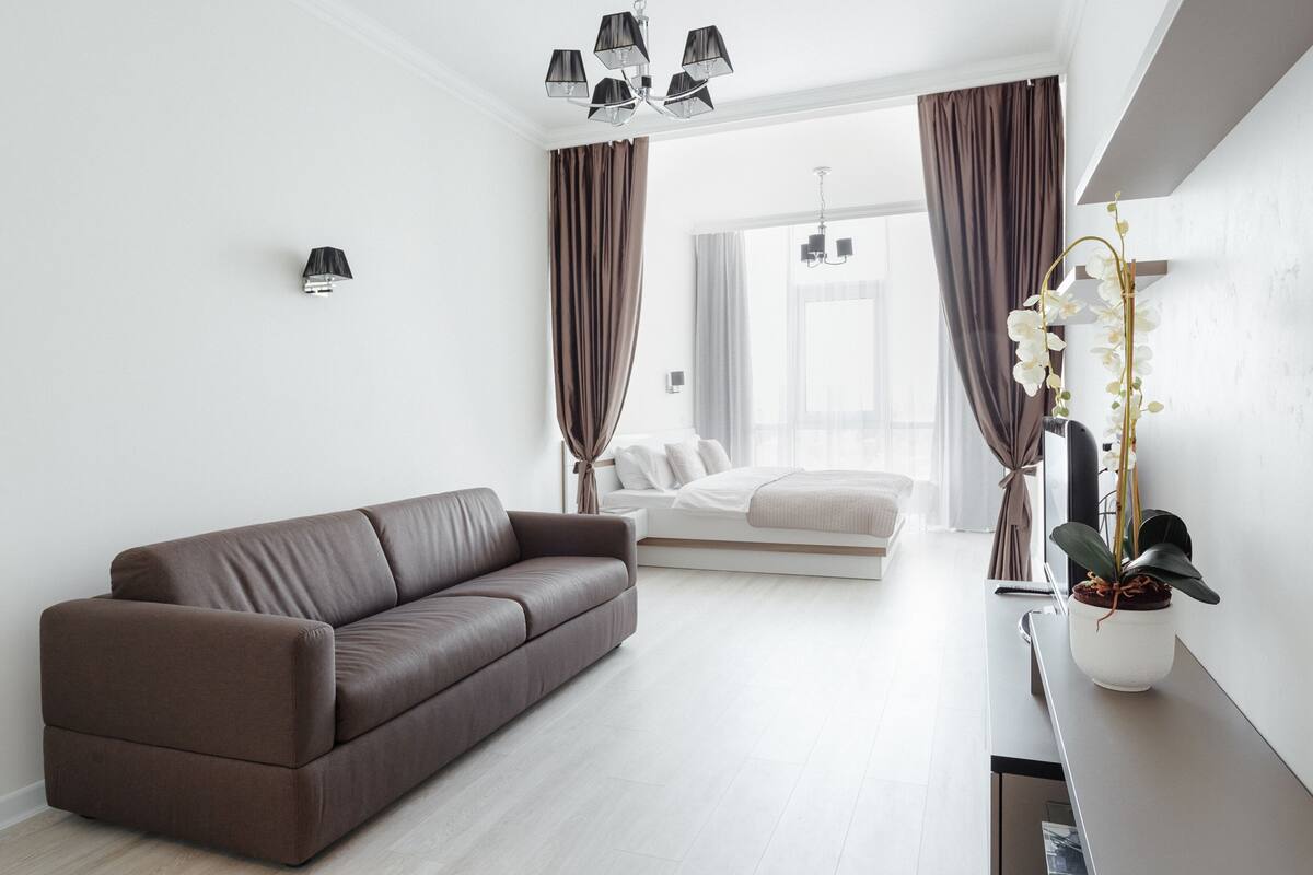 Un dormitorio moderno con un sofá marrón, una cama con ropa de cama blanca, un ventanal con cortinas y una planta en una mesita de noche.