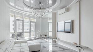 Un salon spacieux doté d’un grand canapé d’angle, d’un lustre moderne, d’un téléviseur à écran plat et offrant une vue sur la ville grâce à de grandes fenêtres.