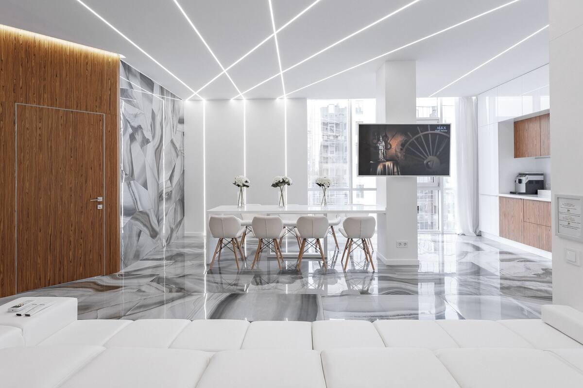 Una cocina moderna con encimera de mármol, una zona de comedor con sillas blancas y un televisor grande en la pared.