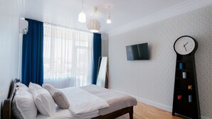 A neatly made bed with white linens and pillows, a wooden headboard, a large window with blue curtains, a wall-mounted TV, and a tall black bookshelf with colorful items. - Royal Apartment Zhukovskogo 9 (Odesa)