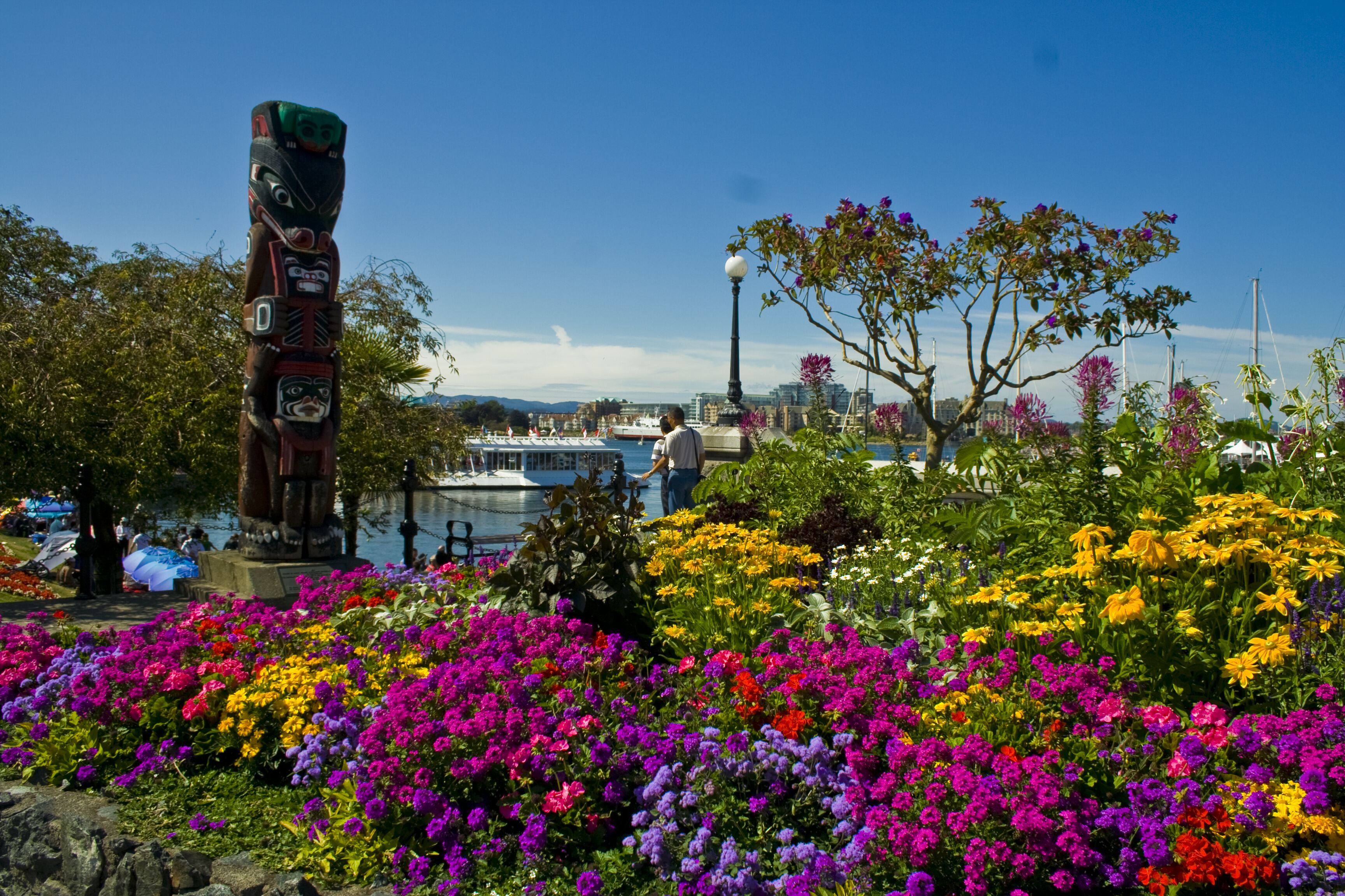 Um totêm e flores coloridas num parque, com um barco e edifícios ao fundo.