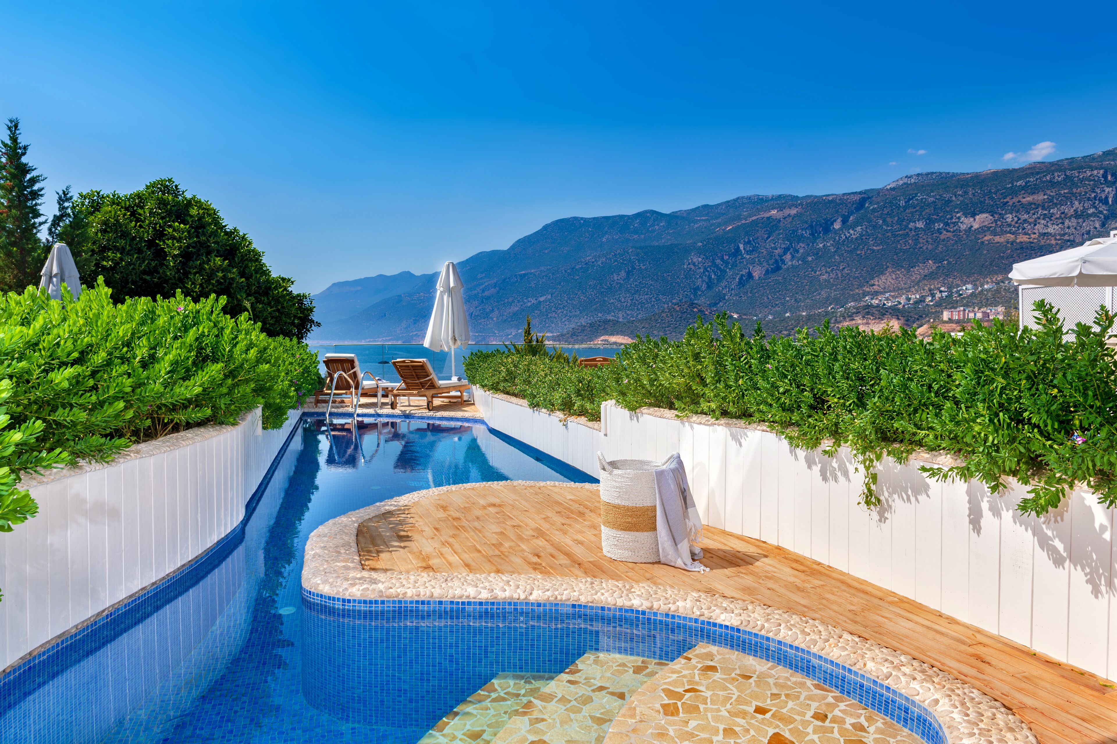 A swimming pool with lounge chairs and umbrellas, surrounded by greenery and mountains in the background.