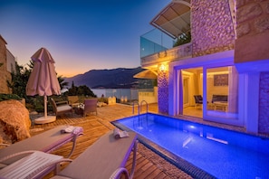 A modern outdoor pool area with lounge chairs, a wooden deck, and a glass balustrade overlooking a body of water. - Peninsula Gardens Hotel (Kas)