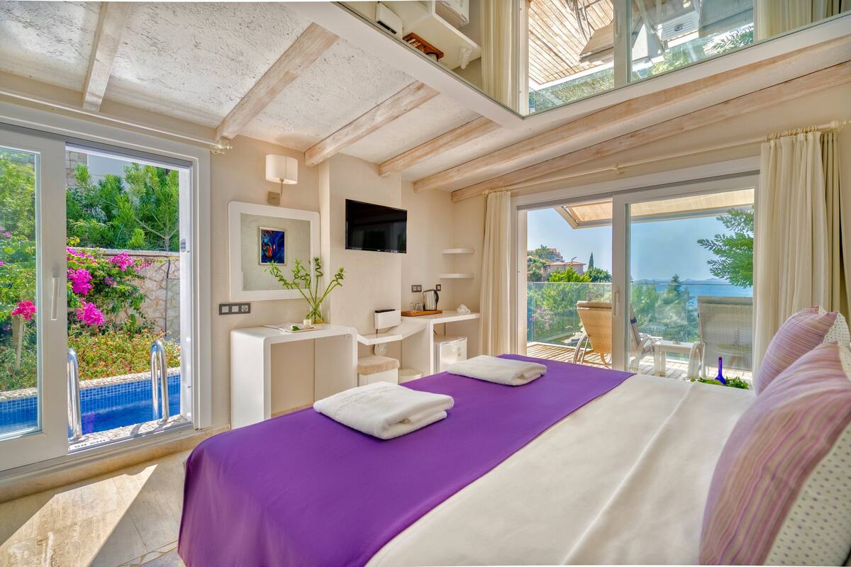 A bedroom with a large bed, a desk, a TV, and a view of a pool and greenery.