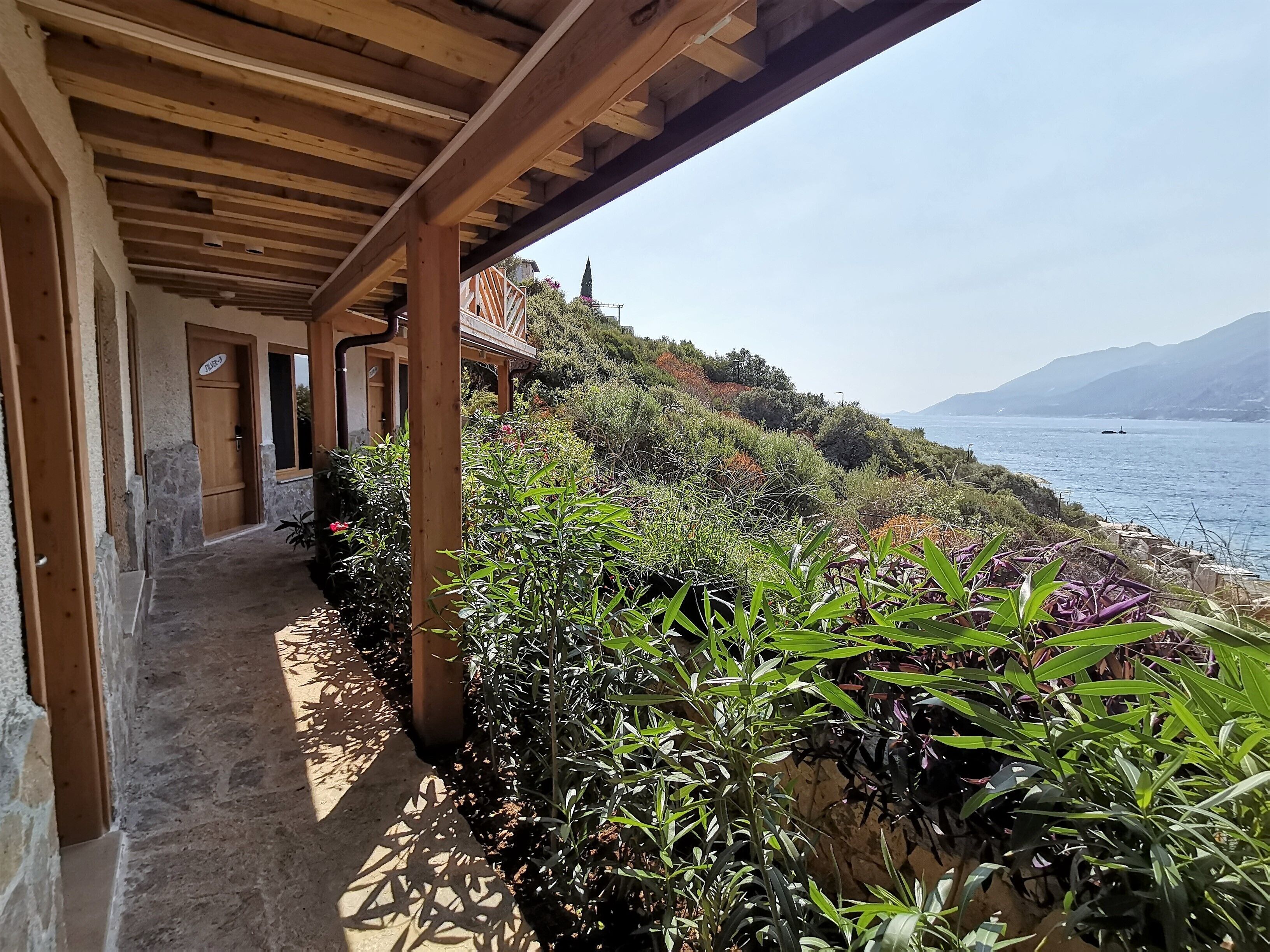 A covered outdoor corridor with stone walls and wooden beams, leading to a scenic coastal view with vegetation and a distant mountain.