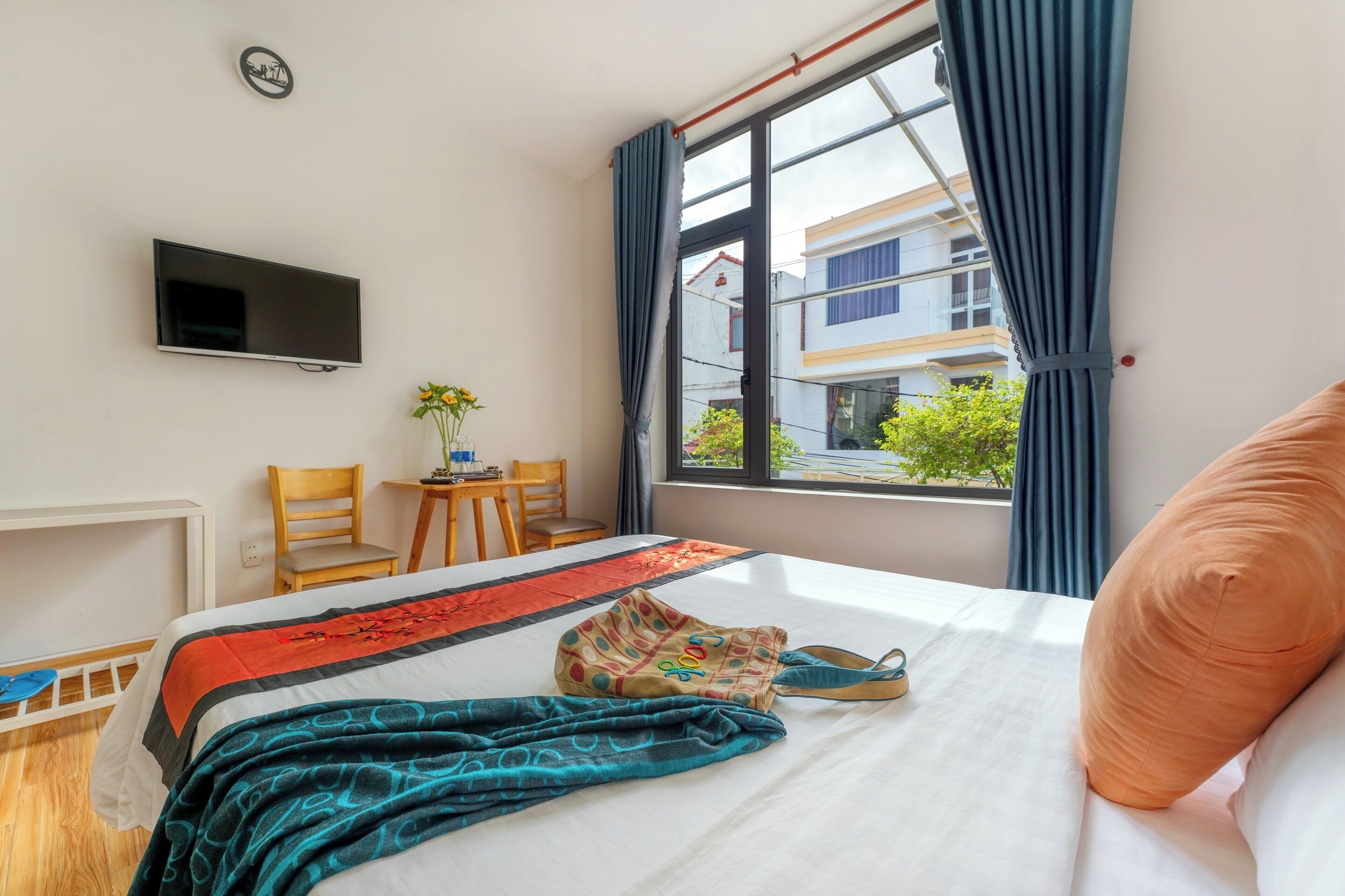 Quarto de hotel com cama, televisão, janela com cortinas, pequena mesa e cadeira, e vista para edifícios.