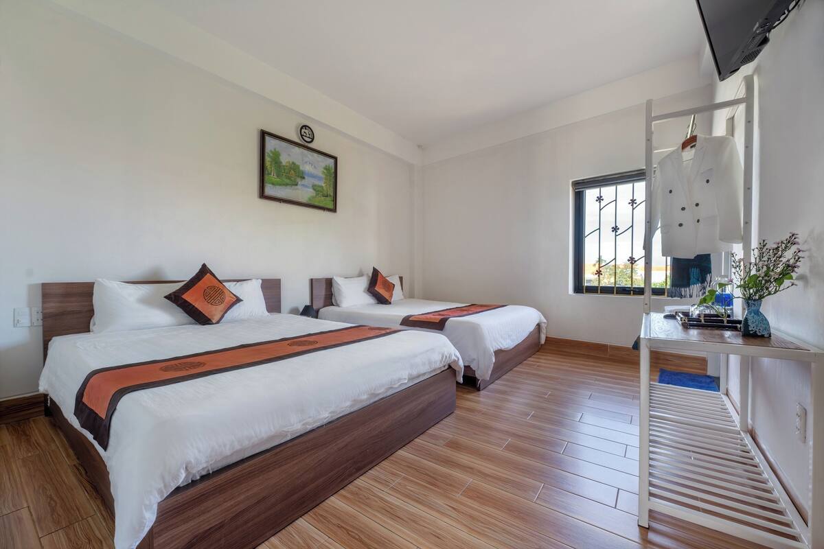 A hotel room with two beds, a wooden floor, a window with a view, a desk with a vase, and a painting on the wall.