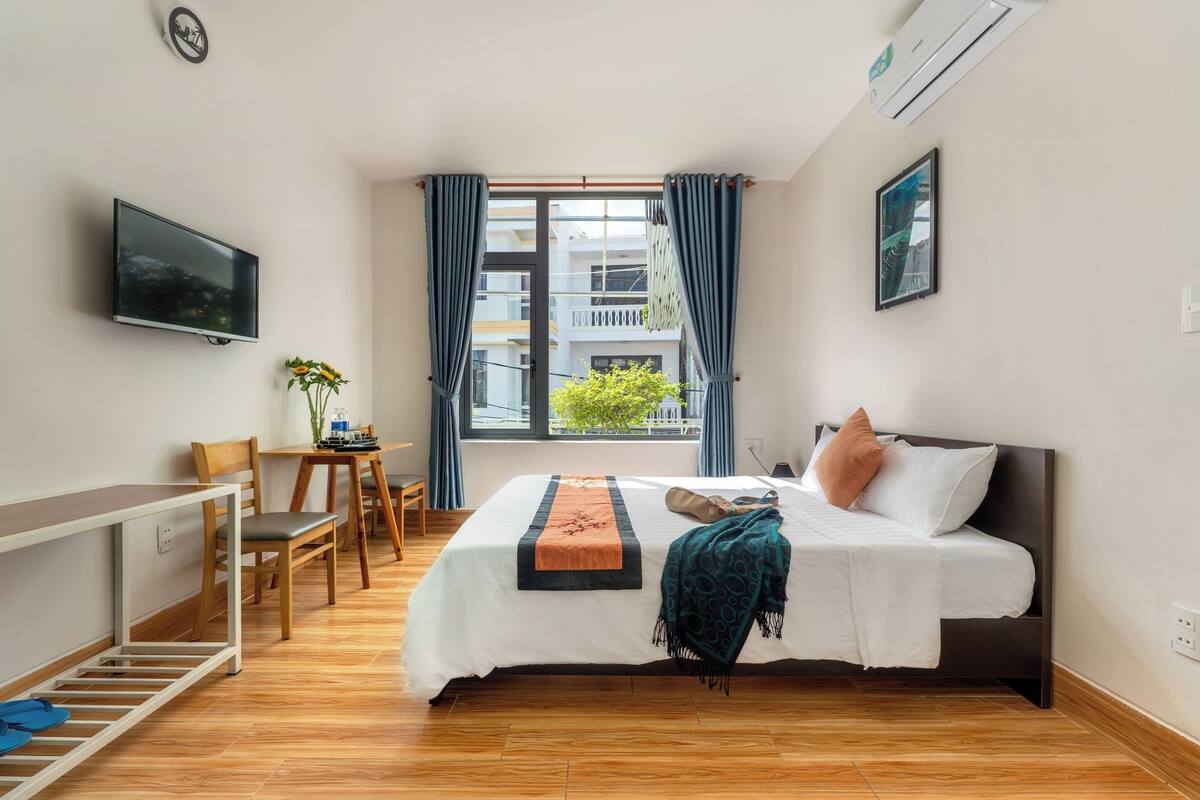 A bedroom with a bed, a desk, a chair, a TV, and a window with curtains.