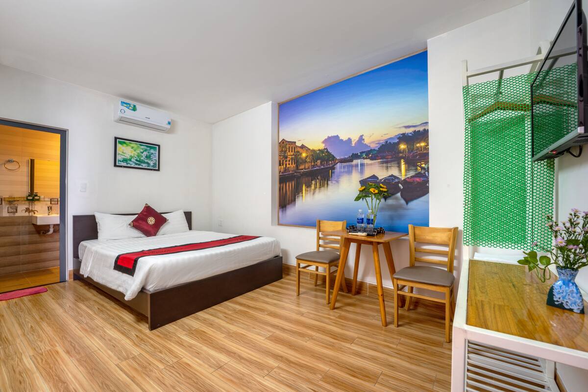 A modern hotel room with a large bed, wooden floor, a desk with two chairs, a painting of a cityscape, and an air conditioning unit.