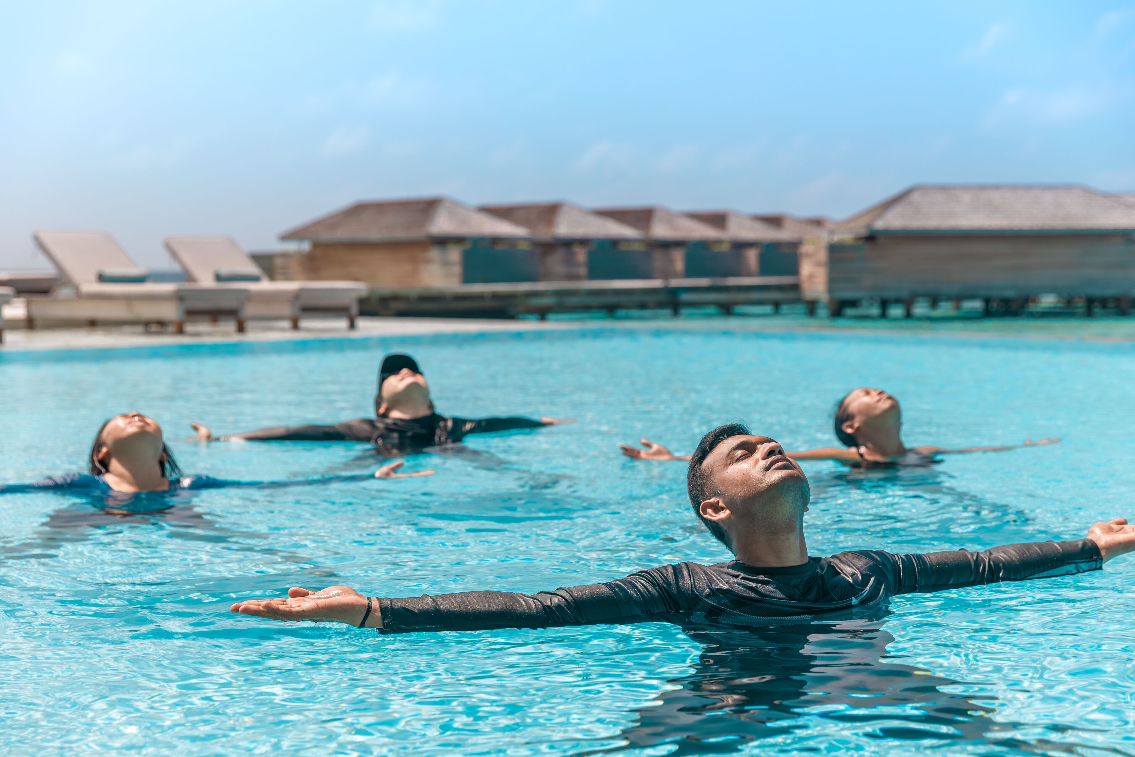 A swimming pool with people in the water, lounge chairs, and overwater bungalows in the background.
