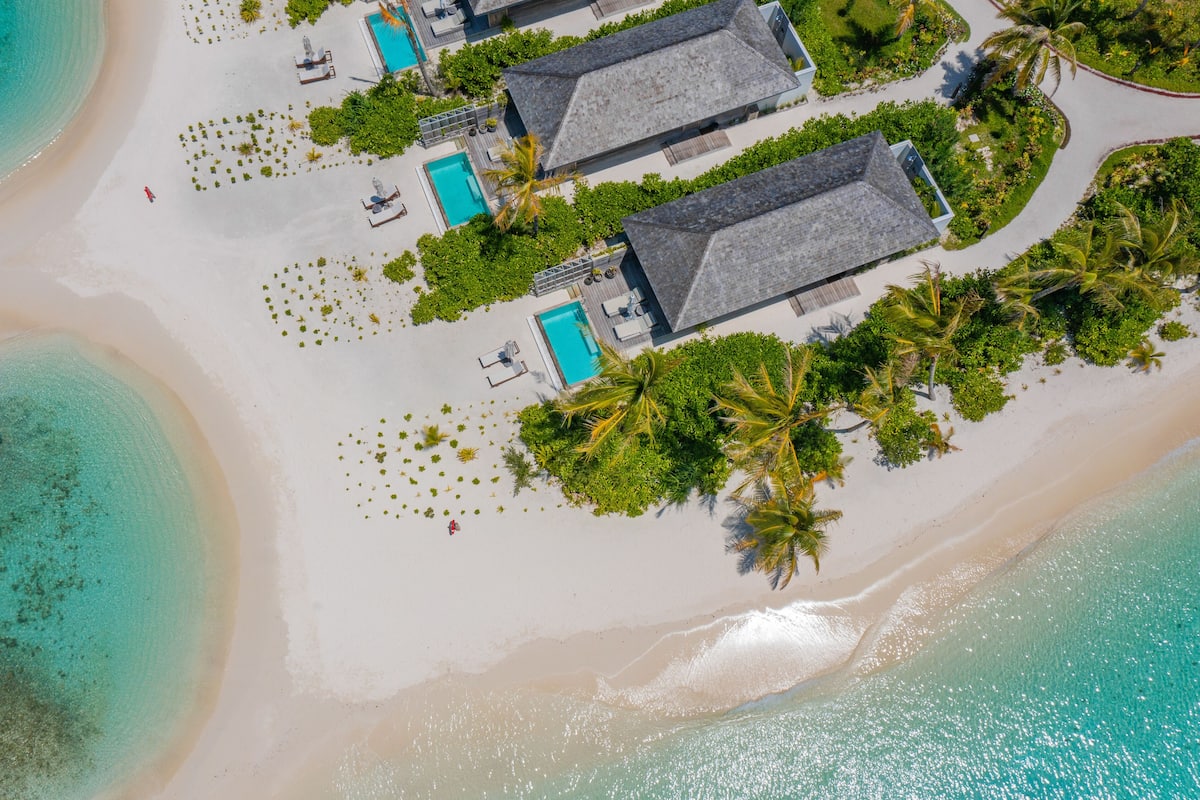 An aerial view of a beachside resort with multiple overwater bungalows, each with a private pool and surrounded by lush greenery and palm trees.