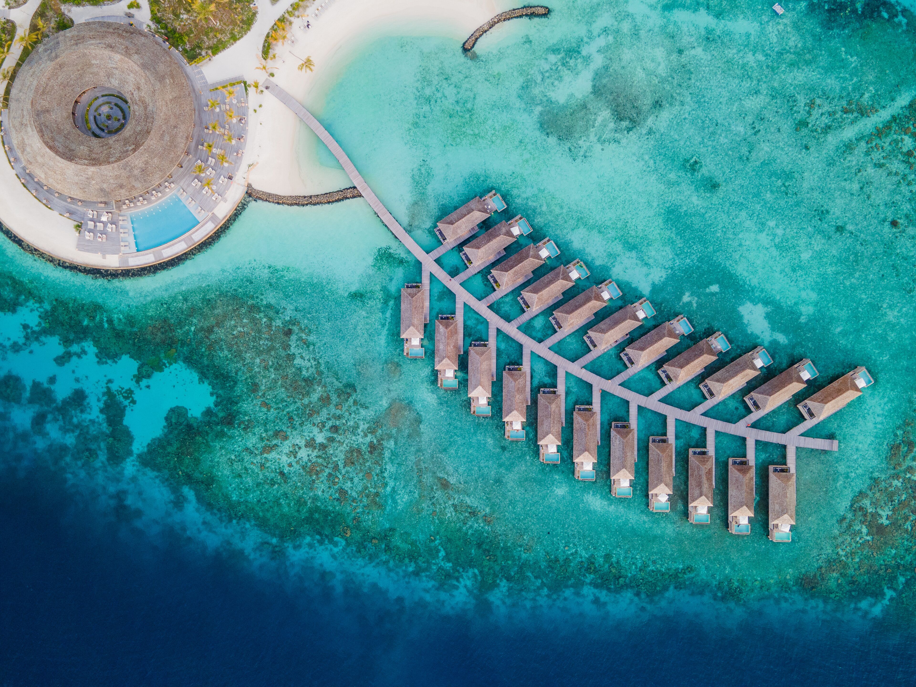 An aerial view of a resort with a central circular building, a crescent-shaped waterway, and a row of overwater bungalows.