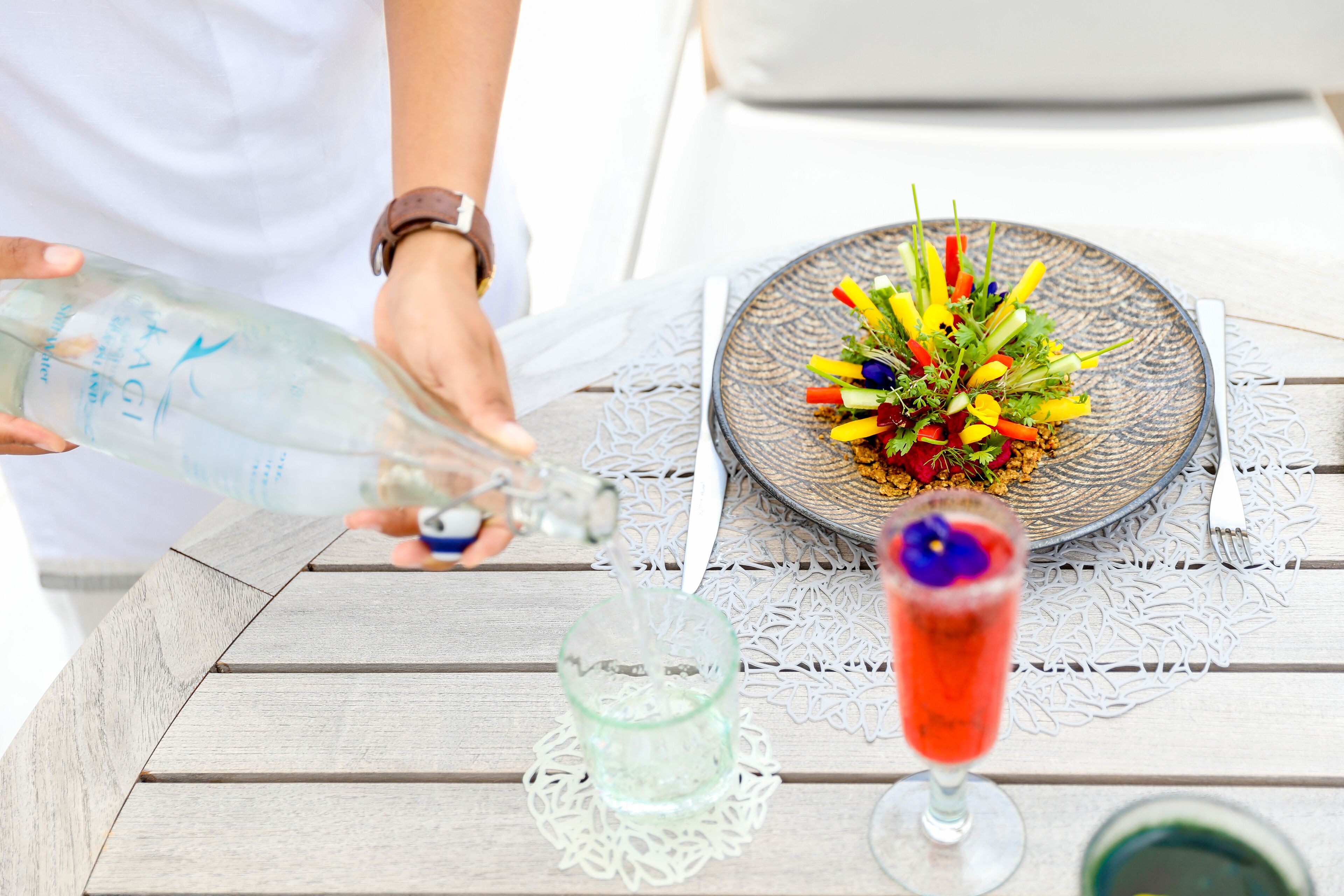 Una mesa con un plato de flores coloridas, un vaso de bebida roja y una botella vertiéndose en un vaso.