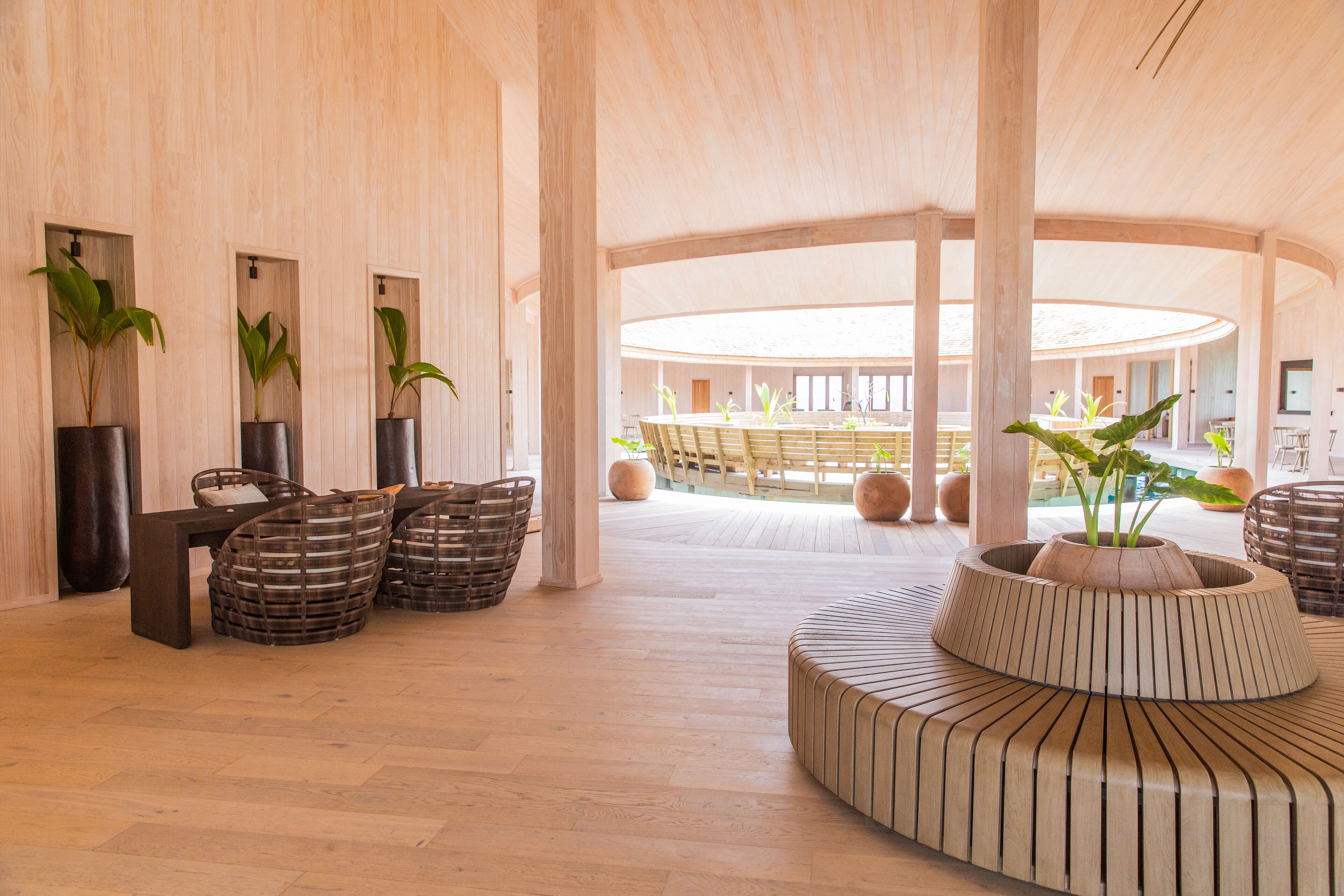 A spacious lobby with wooden flooring, a central seating area with a circular wooden bench, and potted plants.