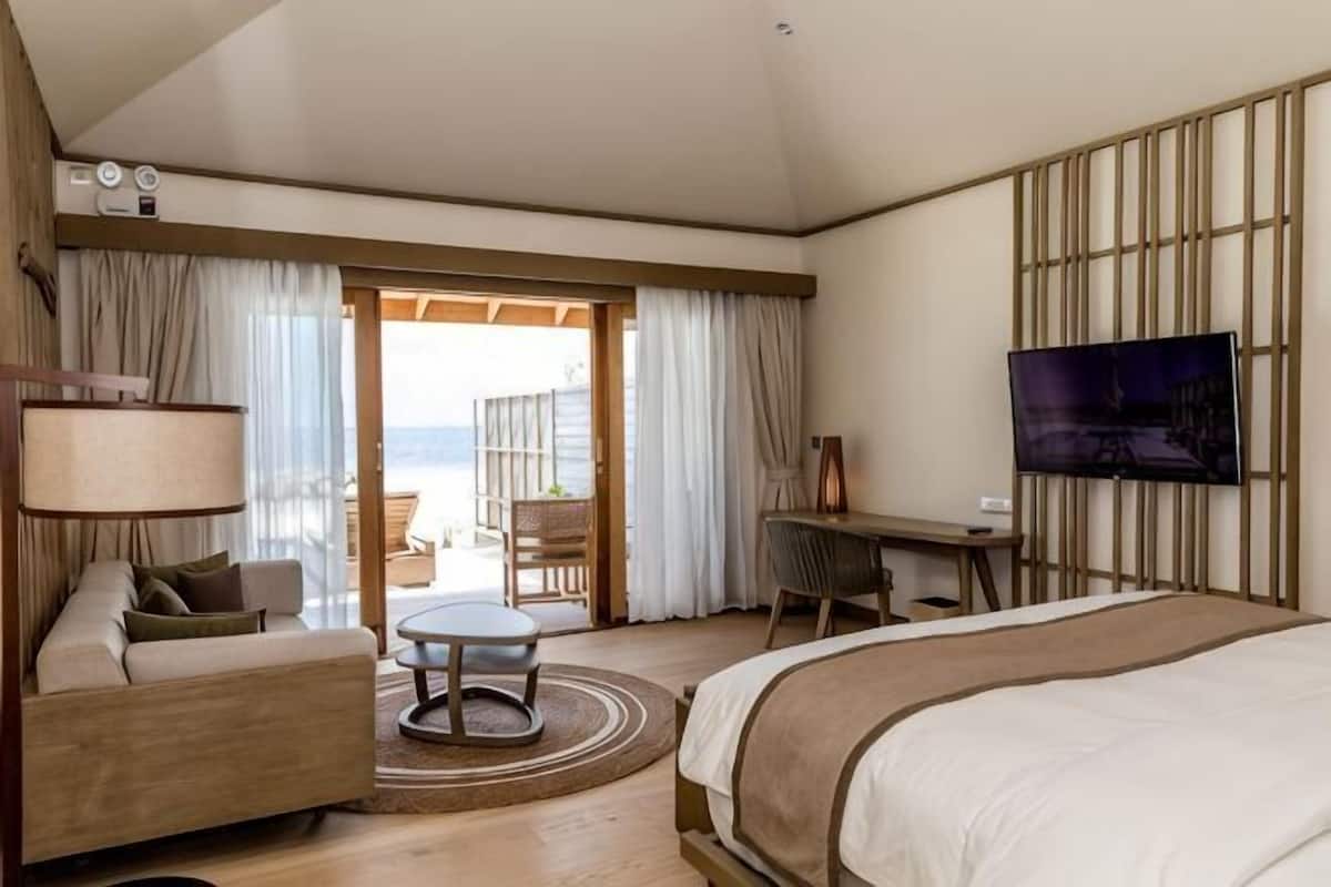 A hotel room with a bed, a desk, a chair, a sofa, a TV, and a view of the beach.
