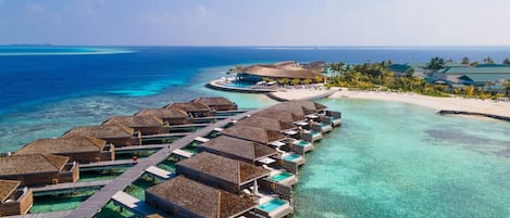 An overwater resort with thatched-roof bungalows connected by walkways, clear turquoise waters, and a beach with palm trees.