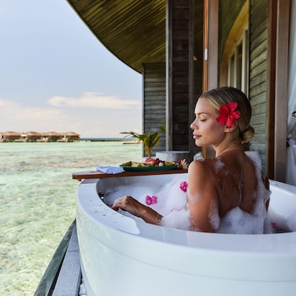 A person relaxing in a bathtub with a view of a beach and overwater bungalows.