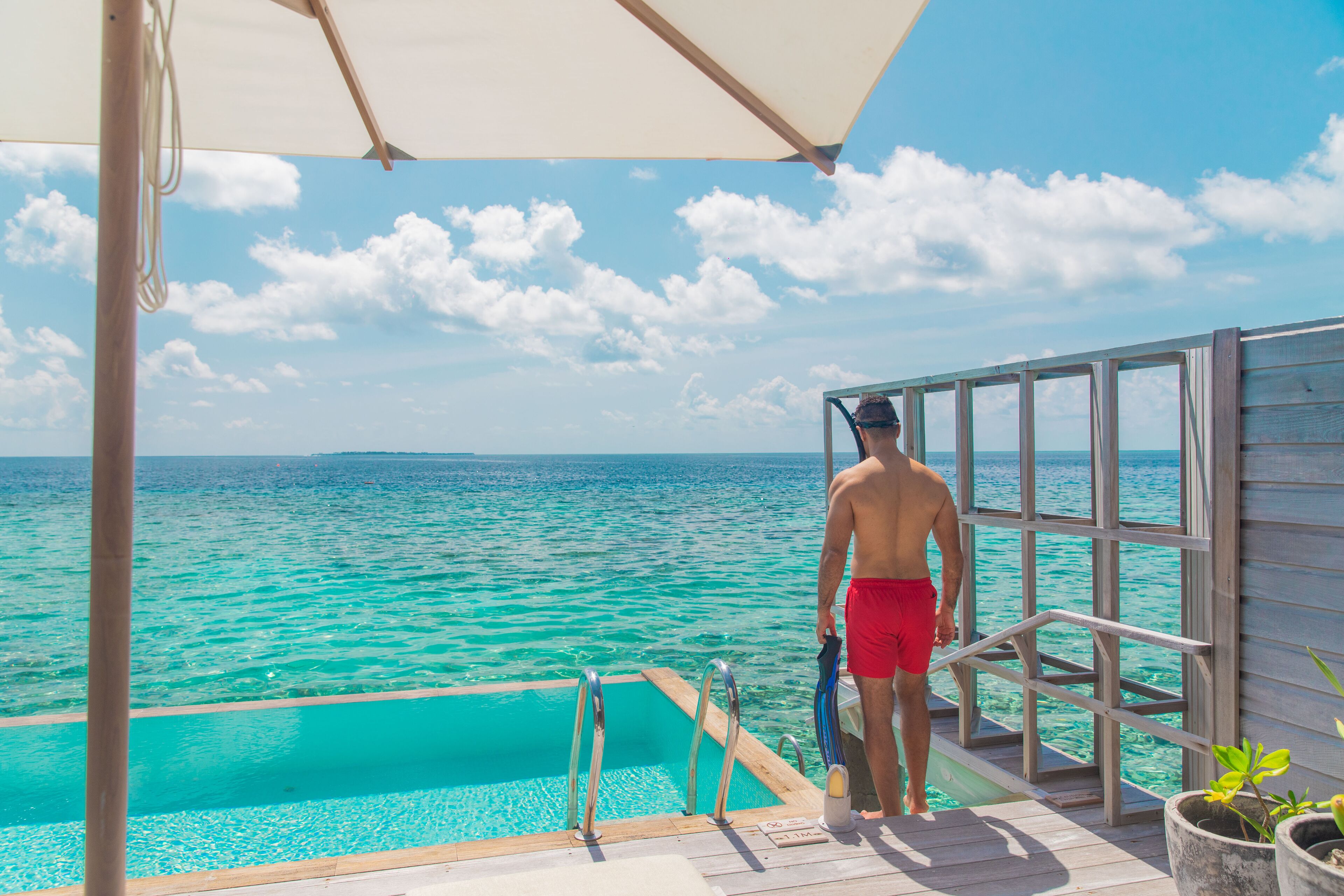 Un hombre con bañador rojo está de pie sobre una terraza de madera, con una piscina y vistas a aguas turquesas.