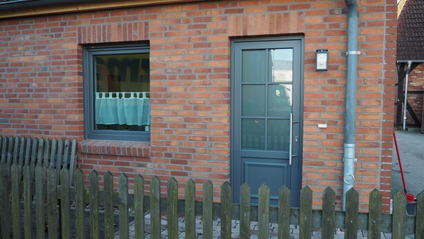 En murstensbygning med en grå dør og et vindue med gardin.